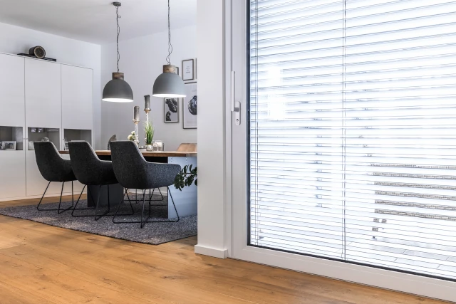 Große, bodentiefe WIRUS Hebeschiebetür aus Glas mit geschlossenem Lamellen-Rollo und prominentem, abschließbarem Griff im Vordergrund; dahinter ein moderner Essbereich mit Tisch, Stühlen und Hängelampen.