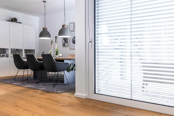Moderner Essbereich mit Holzboden, Esstisch und gepolsterten Stühlen unter zwei Hängelampen; rechts eine große Glas-Schiebetür mit geschlossenen Jalousien.